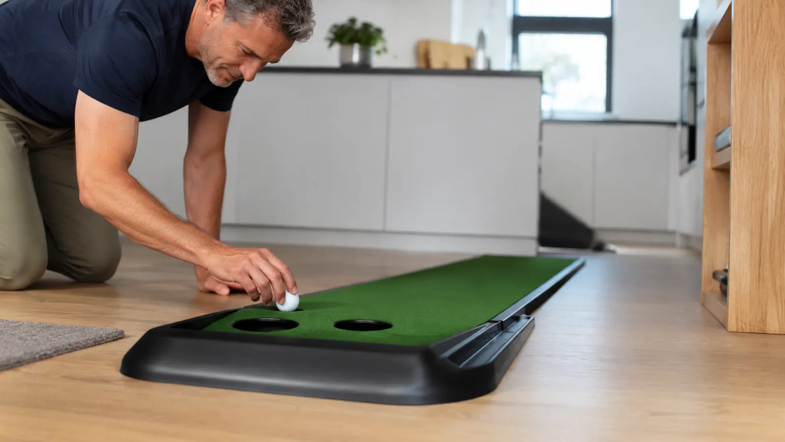 A hand placing a golf ball on a home putting mat with the ball return rail beside it